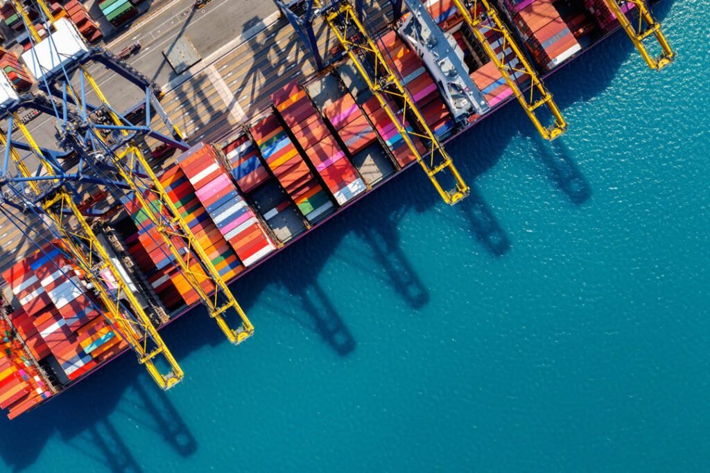 Vista aérea de porto com guindastes e contêineres coloridos atracados sobre o mar azul.